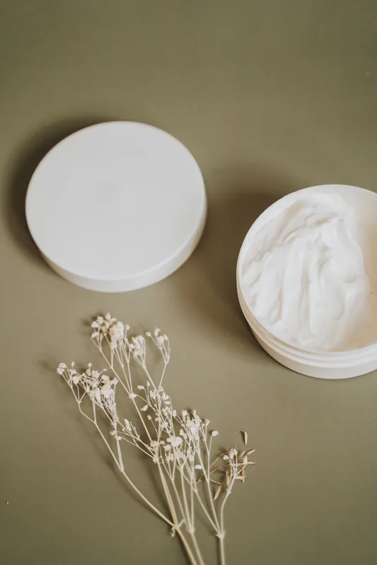 Open jar of white cream beside dried flowers. Illustrative stock photograph, not the product.