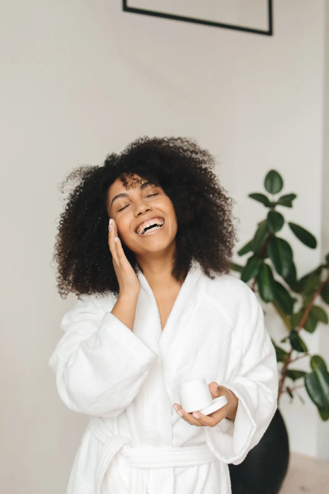 Woman in a bathrobe laughing as she applies cream, holding a plain jar. Illustrative stock photograph, not a patient.