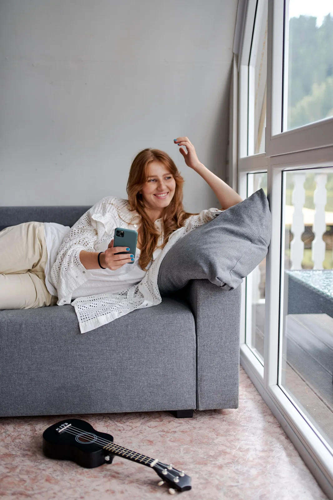 Woman on her sofa checking her phone by the window. Illustrative stock photograph, not a patient.